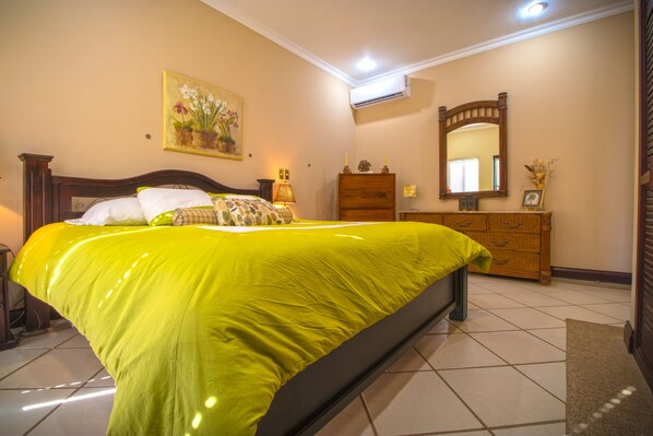 A bedroom with a large bed, a dresser, a mirror, and a painting on the wall. - Visit Sunset Village at Costa Rica, your lovely ocean front place. (Hacienda Pinilla)