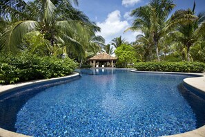 A clear blue pool with a gazebo in the center, surrounded by lush greenery and palm trees. - Visit Sunset Village at Costa Rica, your lovely ocean front place. (Hacienda Pinilla)