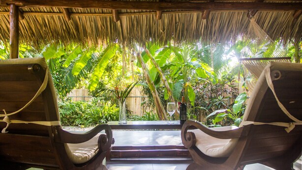 Two wooden lounge chairs with cushions, a glass of wine, and a bottle on a table under a thatched roof, surrounded by lush tropical plants. - Honeymoon Bungalow in Banana Beach with AC, High Speed Wifi, Kitchen, Patio (Cobano)