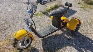 Un scooter jaune est garé sur une plage de galets, avec un banc et une côte rocheuse en arrière-plan.