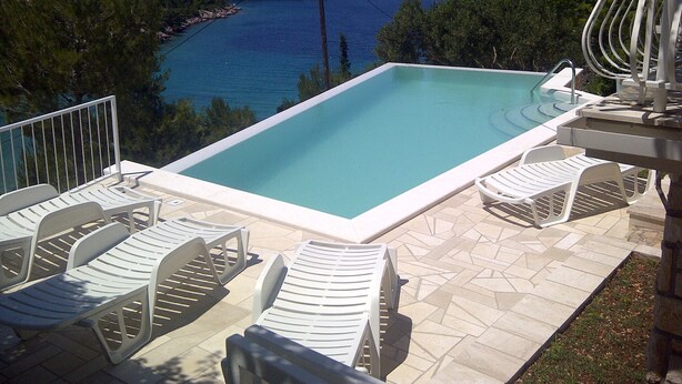 Une piscine rectangulaire entourée d’une terrasse carrelée, avec des chaises blanches et une table.