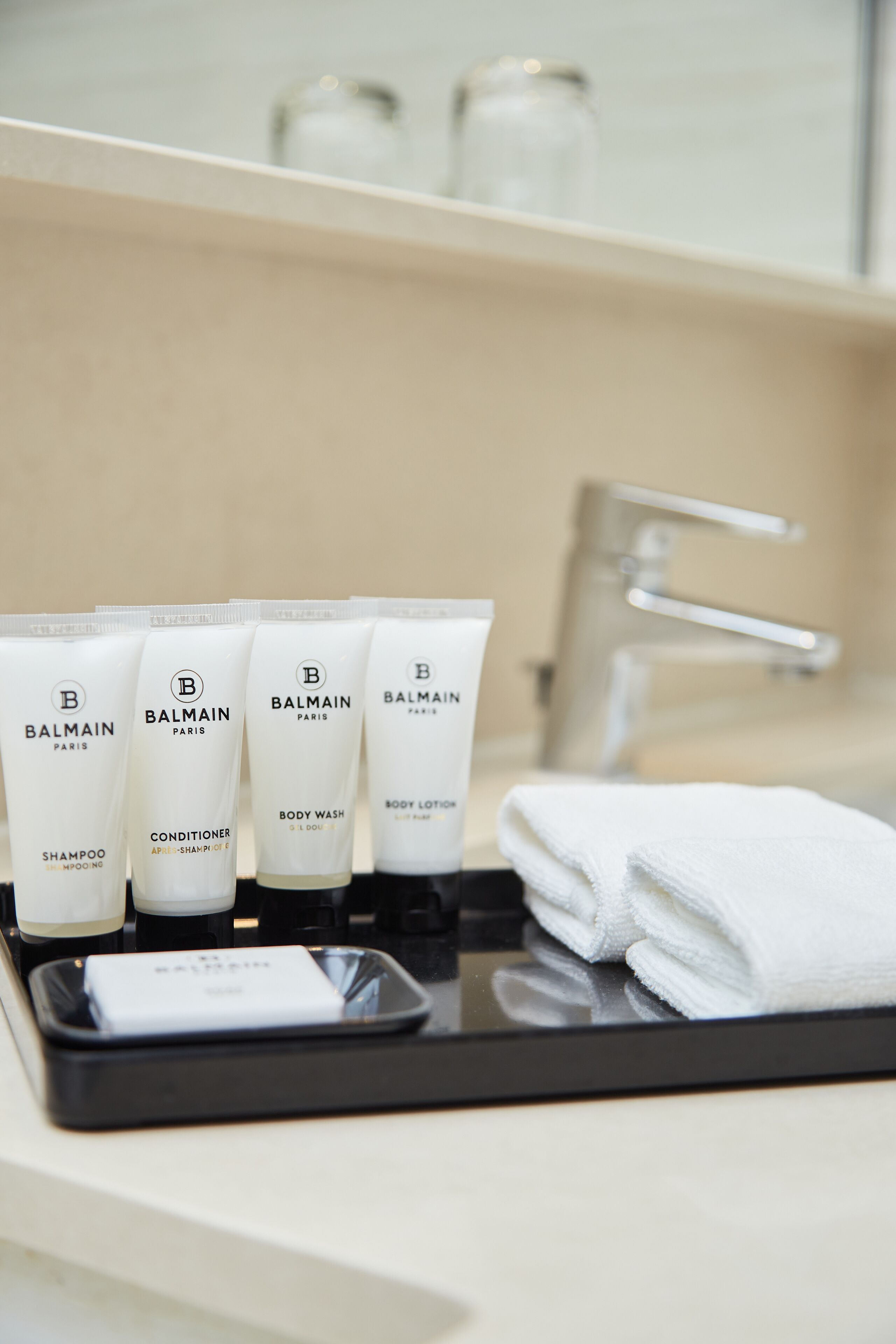 Ein Badezimmer-Waschtisch mit einer Ablage, auf der sich vier Flaschen Balmain Paris Duschgel und Conditioner, ein gefaltetes weißes Handtuch und ein Smartphone befinden.