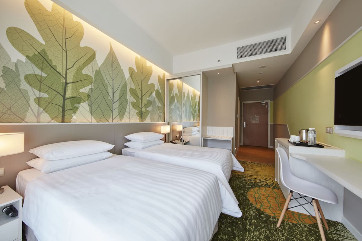 Hotelkamer met twee bedden, een bureau, een stoel, een wand met groot bladmotief en een kleine kitchenette.