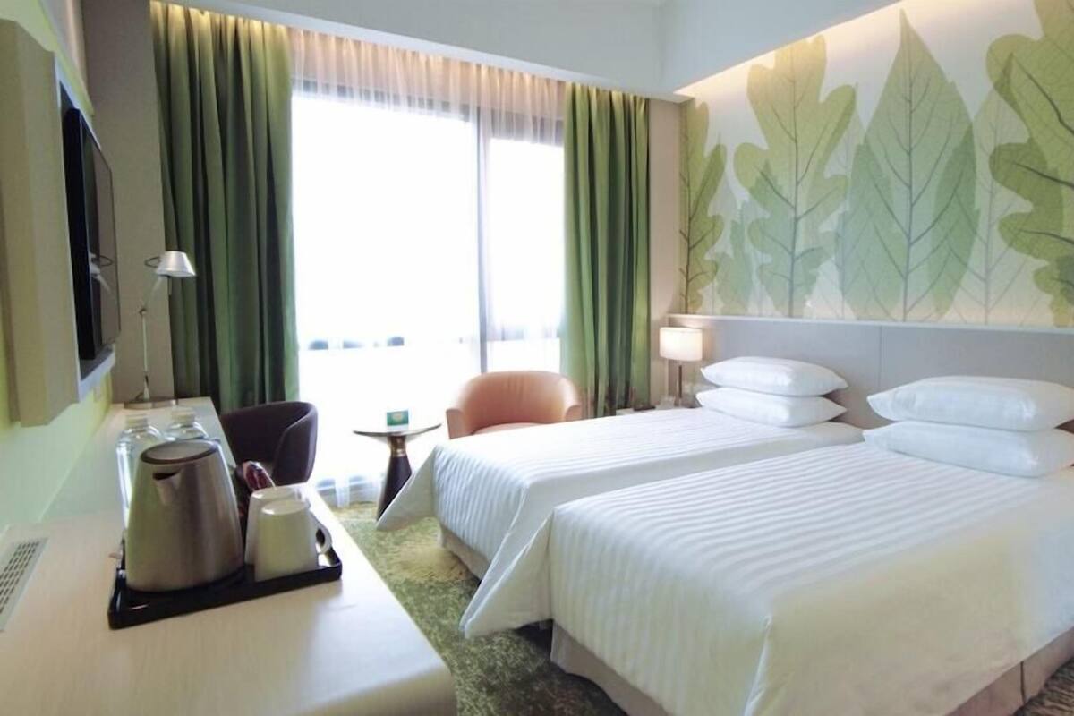 Een hotelkamer met twee bedden, een bureau, een stoel, een koffiezetapparaat en een grote wand met een bladmotief.