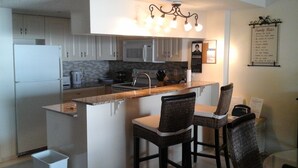 A modern kitchen with white cabinets, a granite countertop, and a stainless steel refrigerator. There are two bar stools and a small trash bin. - Beautiful Beachfront Getaway retreat for couples, Private Balcony & Sunsets #202 (Indian Shores)