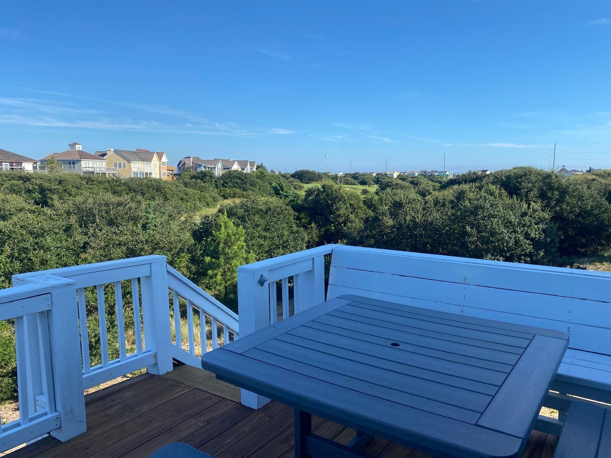Um deck de madeira com guarda-corpo branco e uma mesa, com vista para uma paisagem verdejante e casas ao longe.