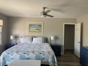 Un dormitorio con una cama, dos mesitas de noche, un ventilador de techo, una puerta y un cuadro enmarcado en la pared.