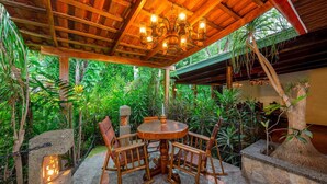 A wooden outdoor seating area with a round table and chairs, surrounded by lush greenery and a wooden ceiling with hanging lights. - Escazu Vacation  Rentals, Studio Apartments and standard rooms (San Jose)