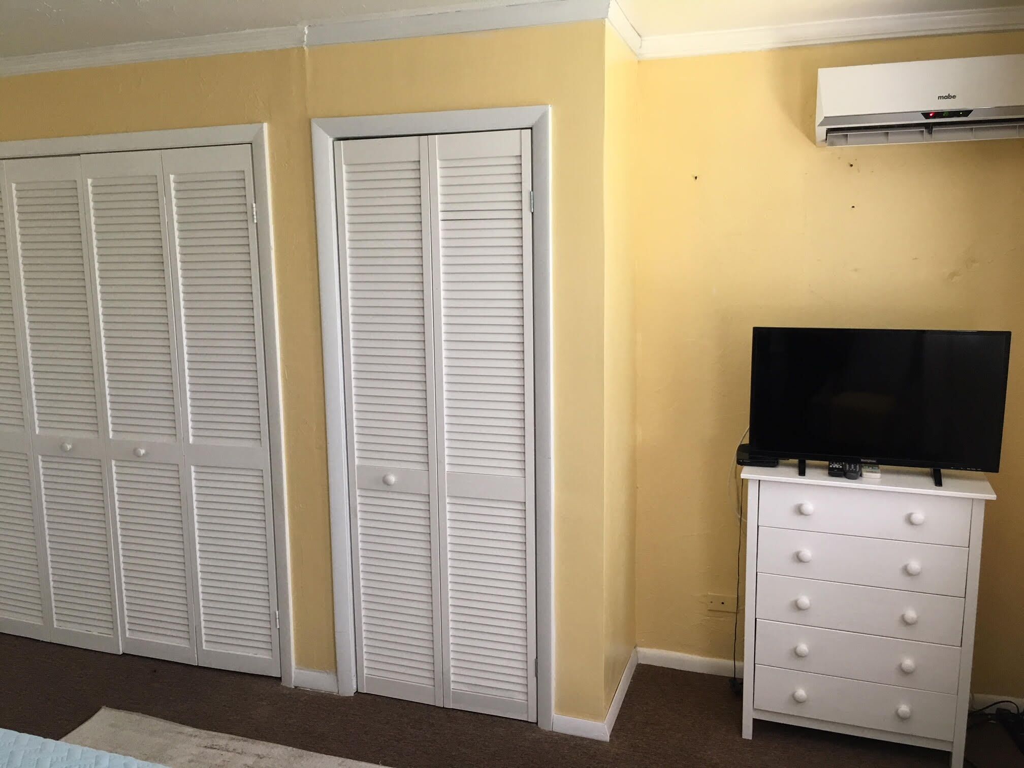 Una habitación con dos armarios blancos con puertas a persiana y un tocador blanco con cajones, además de un aire acondicionado instalado en la pared.