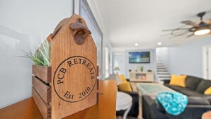 Une enseigne en bois portant l’inscription « PCB RETREATS Fondé en 2010 » est posée sur une étagère en bois dans un salon moderne, avec un canapé et un téléviseur en arrière-plan.