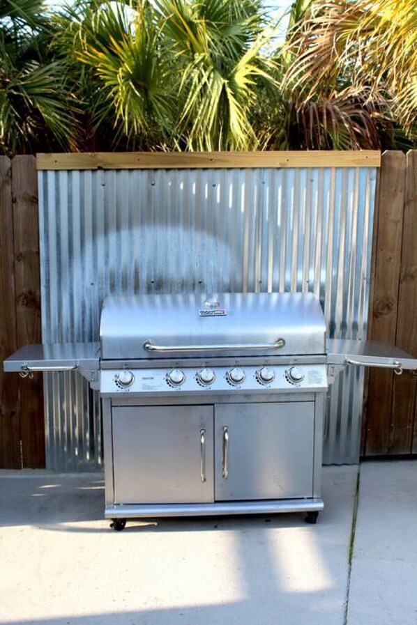 Una parrilla de acero inoxidable con cuatro quemadores y una tapa, ubicada al aire libre con una cerca de madera y palmeras al fondo.
