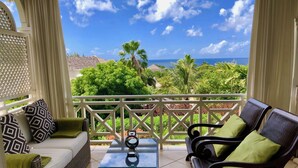 Un balcon avec vue sur l’océan, un canapé en osier avec des coussins verts, une petite table agrémentée d’un objet décoratif et une balustrade blanche.