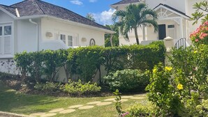 Un bâtiment blanc surmonté d’un toit en tuiles, entouré d’une végétation luxuriante et d’un jardin bien entretenu, agrémenté de pierres plates servant de pas.