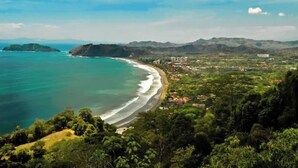 A coastal landscape with a beach, forested hills, and a distant cityscape. - Oceanfront  Beachfront Condo Vacational Rental Jacó Beach Playa Jacó, costa rica (Garabito)