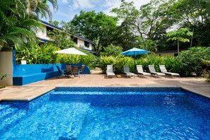 A swimming pool with lounge chairs and umbrellas, surrounded by lush greenery and a building with a blue roof.