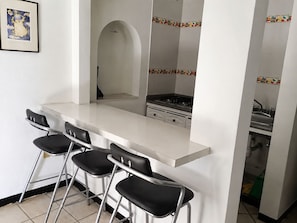A kitchen with a white countertop, four black bar stools, a stove, and a tiled backsplash with fruit patterns.