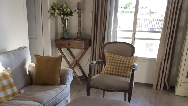 Un salon comprenant un canapé, un pouf et un fauteuil, une petite table avec un vase, et une vue par une fenêtre avec des rideaux.