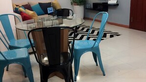 Un coin repas avec une table en verre et quatre chaises, un canapé avec des coussins, une plante en pot et un téléviseur fixé au mur.