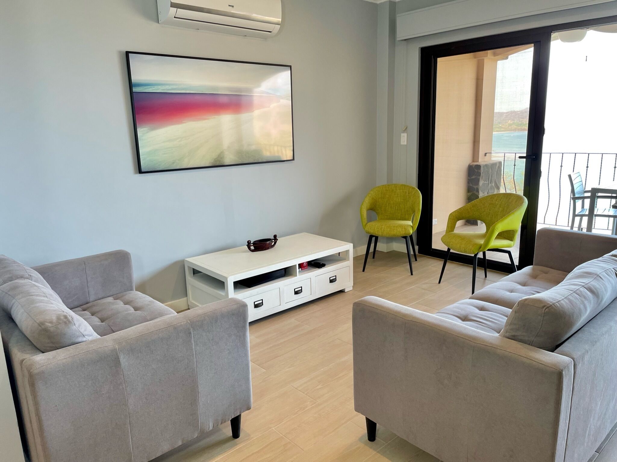 A modern living room with two grey sofas, a white coffee table, and two yellow chairs. There is a large abstract painting on the wall, and a balcony with a view of the sea is visible through the glass door.