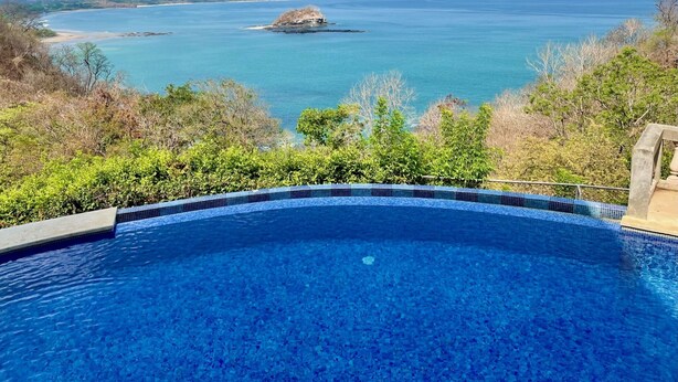 Une piscine d’eau cristalline, d’un bleu éclatant, offrant une vue sur l’océan et les collines lointaines.
