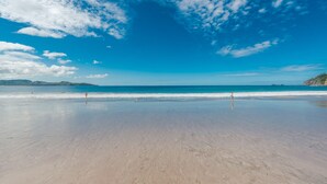 Une vaste plage de sable fin, sous un ciel bleu azur, avec des montagnes qui se dessinent à l’horizon.