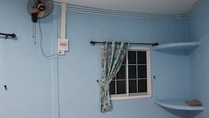A room with light blue walls, a window with floral curtains, a ceiling fan, and a small shelf. - Demy Home (Suan Phueng)