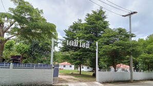 A gated entrance with a sign, surrounded by trees and a white building with a red roof. - Demy Home (Suan Phueng)