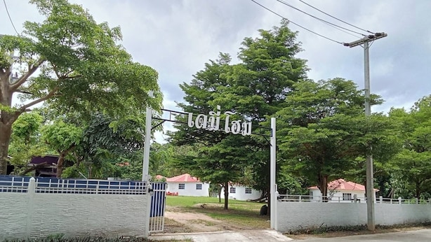 Una entrada protegida por un muro con un letrero, rodeada de árboles y un edificio blanco de techo rojo.