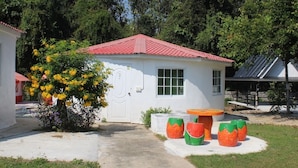 A small white building with a red roof, a door, and windows. There is a tree with yellow flowers and a table with colorful stools. - Demy Home (Suan Phueng)