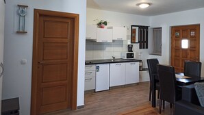 Une cuisine moderne avec des meubles blancs, un four à micro-ondes et une machine à café. Une porte en bois se trouve sur le côté gauche, et un coin repas avec une table et des chaises est aménagé.