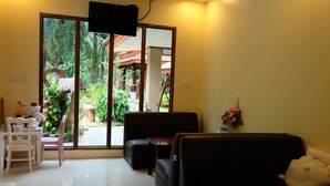 A living room with a sofa, a table, and chairs. There is a TV mounted on the wall, and a window with a view of greenery outside. - Tonpalm Farmstay (Khlong Thom)