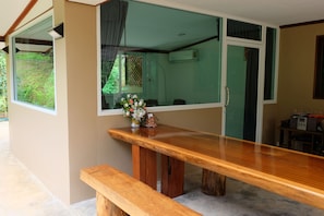 A wooden bench and table in a room with large windows, a small table with a vase of flowers, and a door with a green door frame. - Tonpalm Farmstay (Khlong Thom)