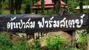 A wooden sign with Thai text in a natural setting with greenery and a red-roofed building in the background. - Tonpalm Farmstay (Khlong Thom)