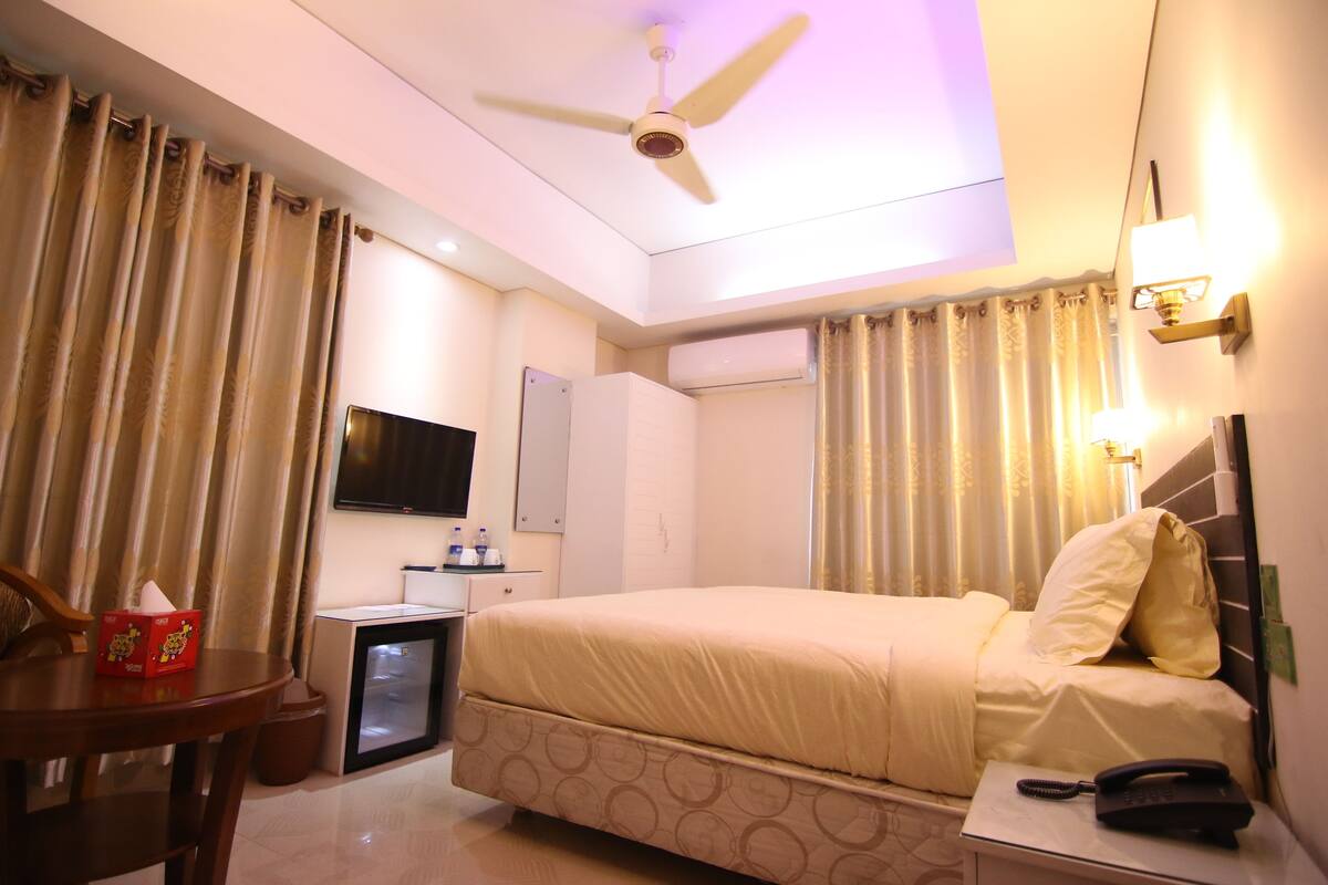 Habitación de hotel con cama, mesita de noche, escritorio, silla, televisor, aire acondicionado y cortinas.