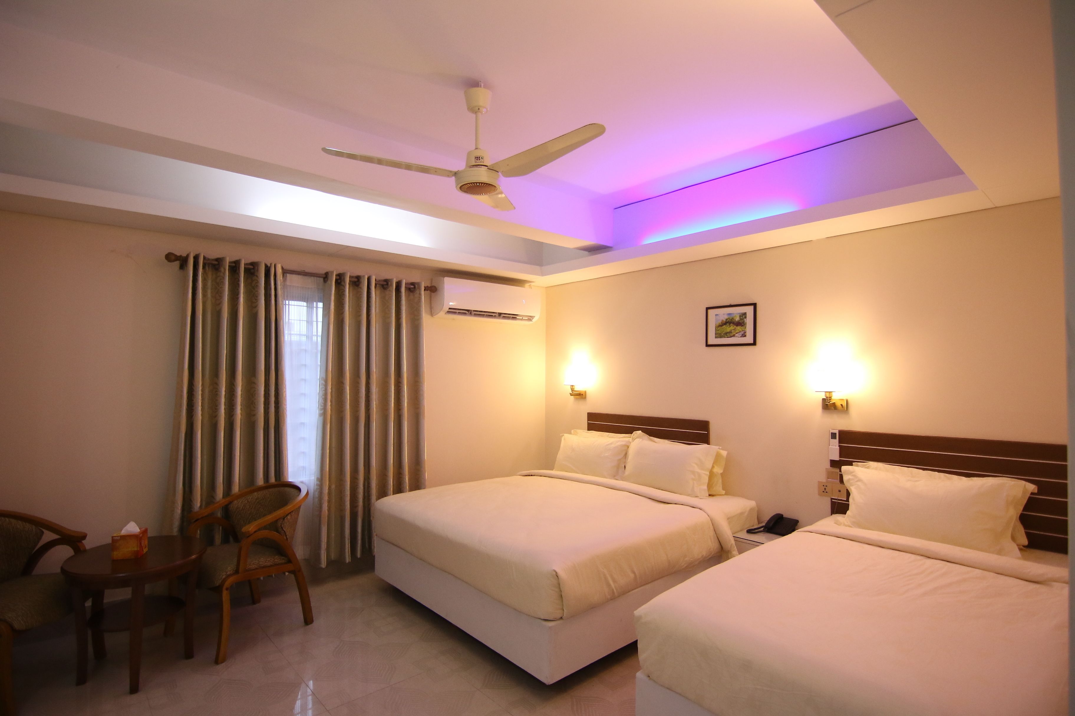 A hotel room with two beds, a ceiling fan, a table with chairs, and a window with curtains.