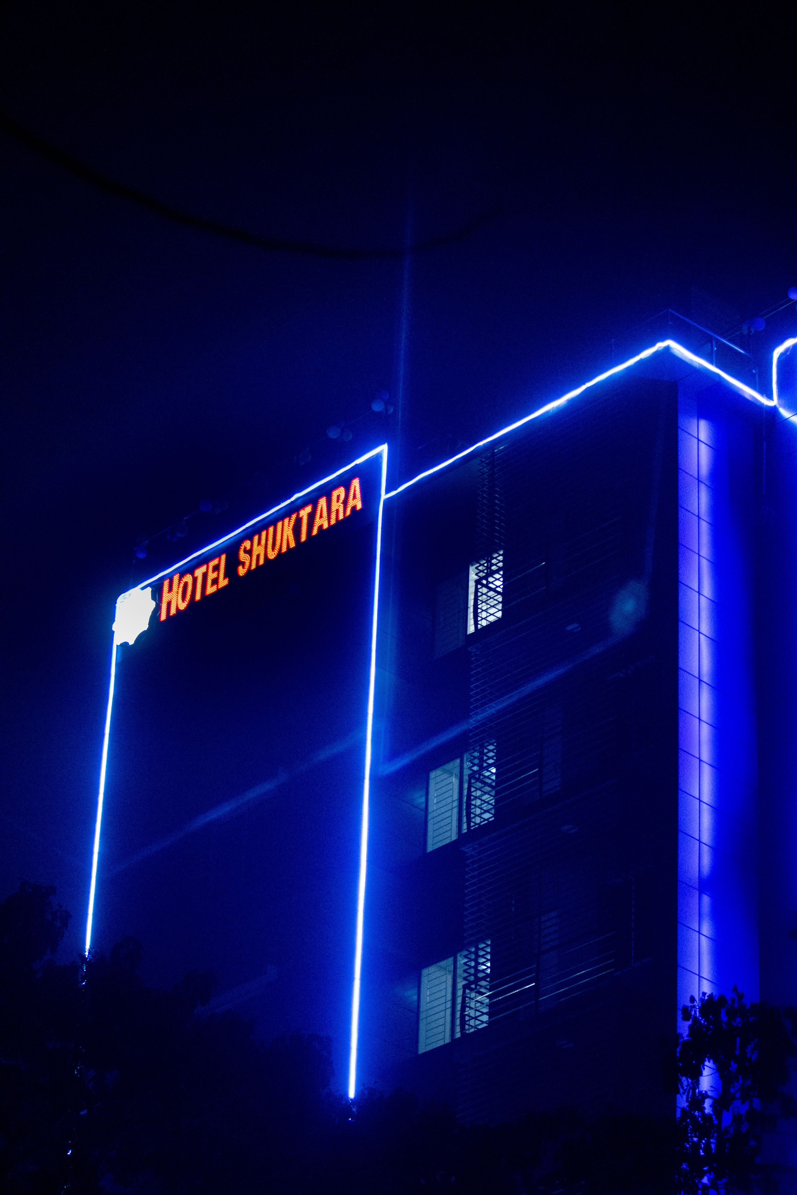 A building with a sign reading "HOTEL SHUKTARA" illuminated by blue neon lights.