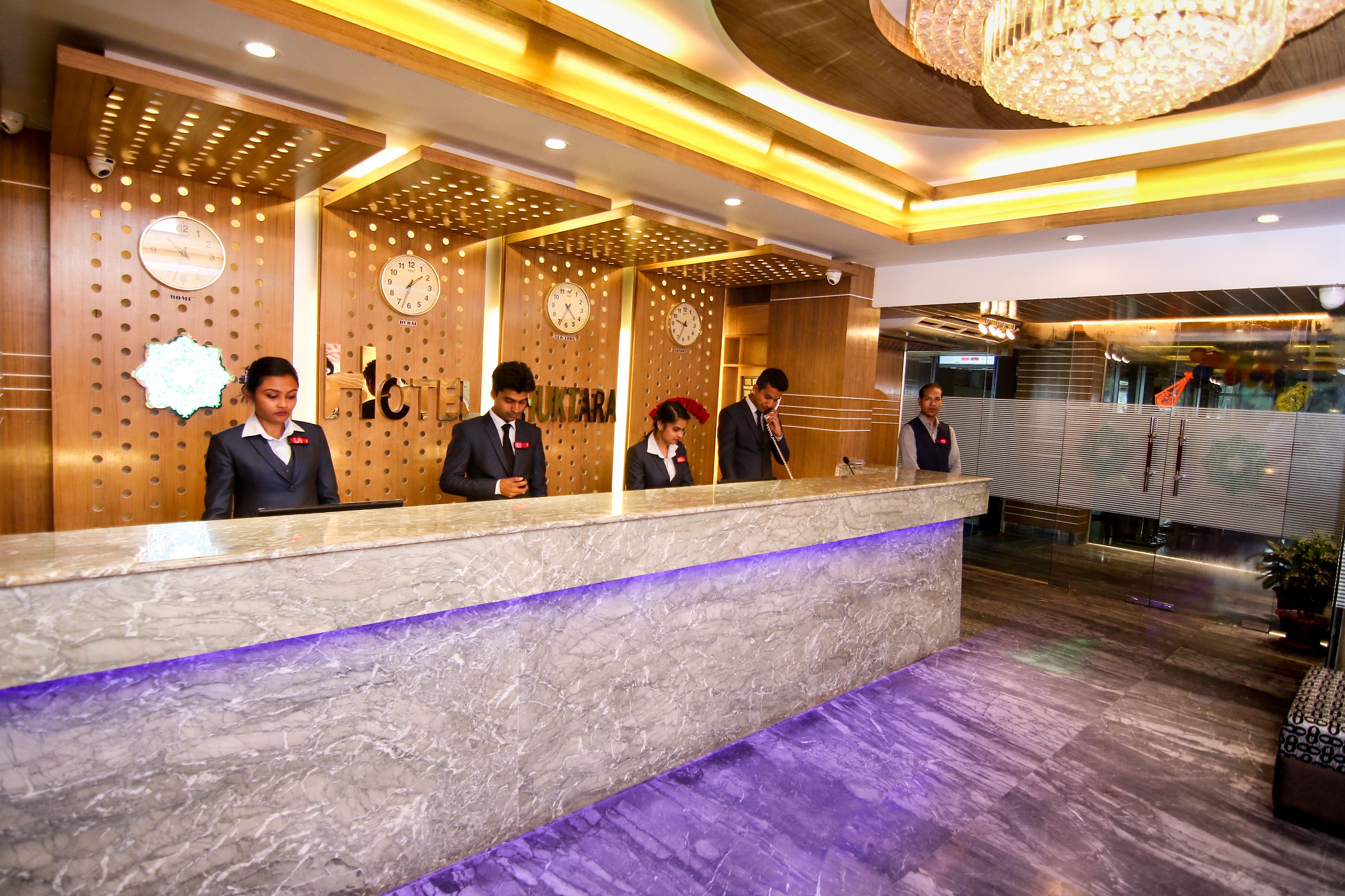 A hotel reception desk with a marble counter, wooden paneling, and clocks on the wall.