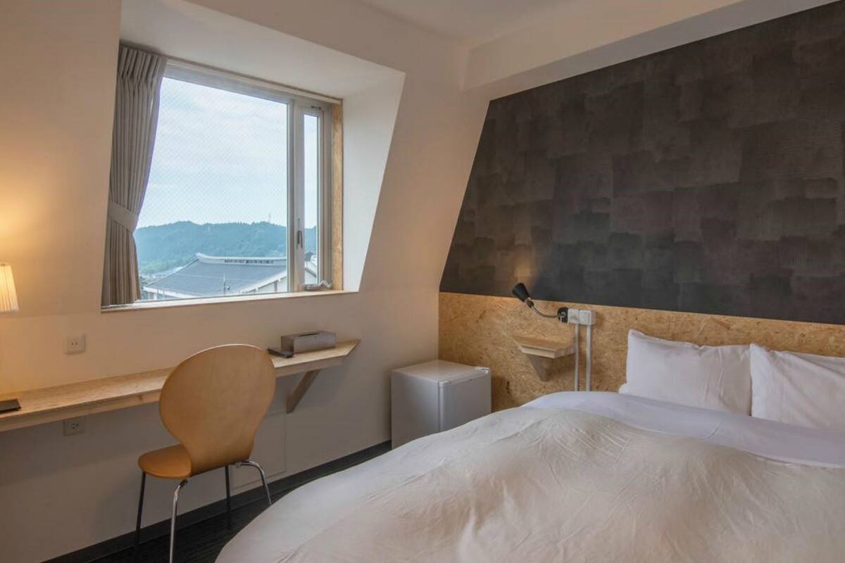 A hotel room with a bed, a desk with a chair, a window with a view, and a wall-mounted lamp.
