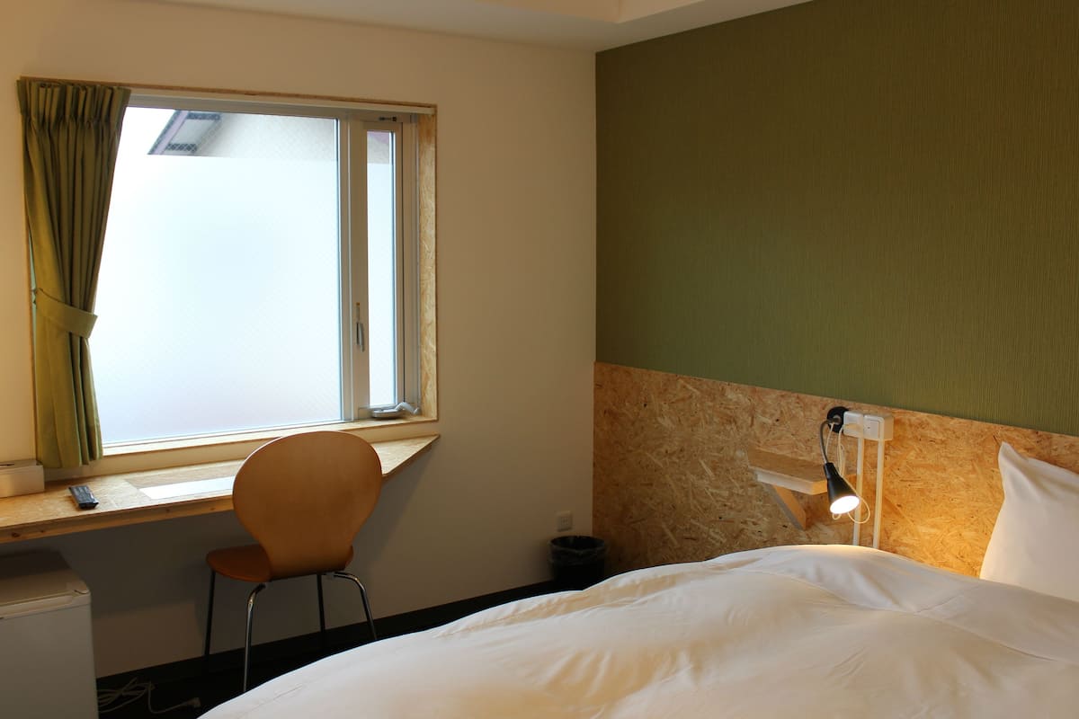 A hotel room with a bed, a desk with a chair, a window with curtains, a wall-mounted lamp, and a small trash bin.