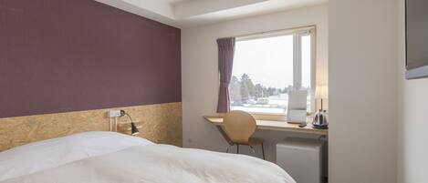 A hotel room with a bed, a desk with a chair, a window with curtains, and a view of trees.