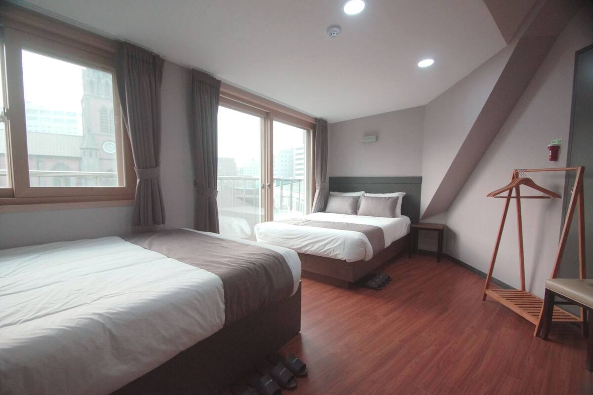 A hotel room with two beds, a wooden floor, a window with curtains, a bedside table, and a hanging rack.
