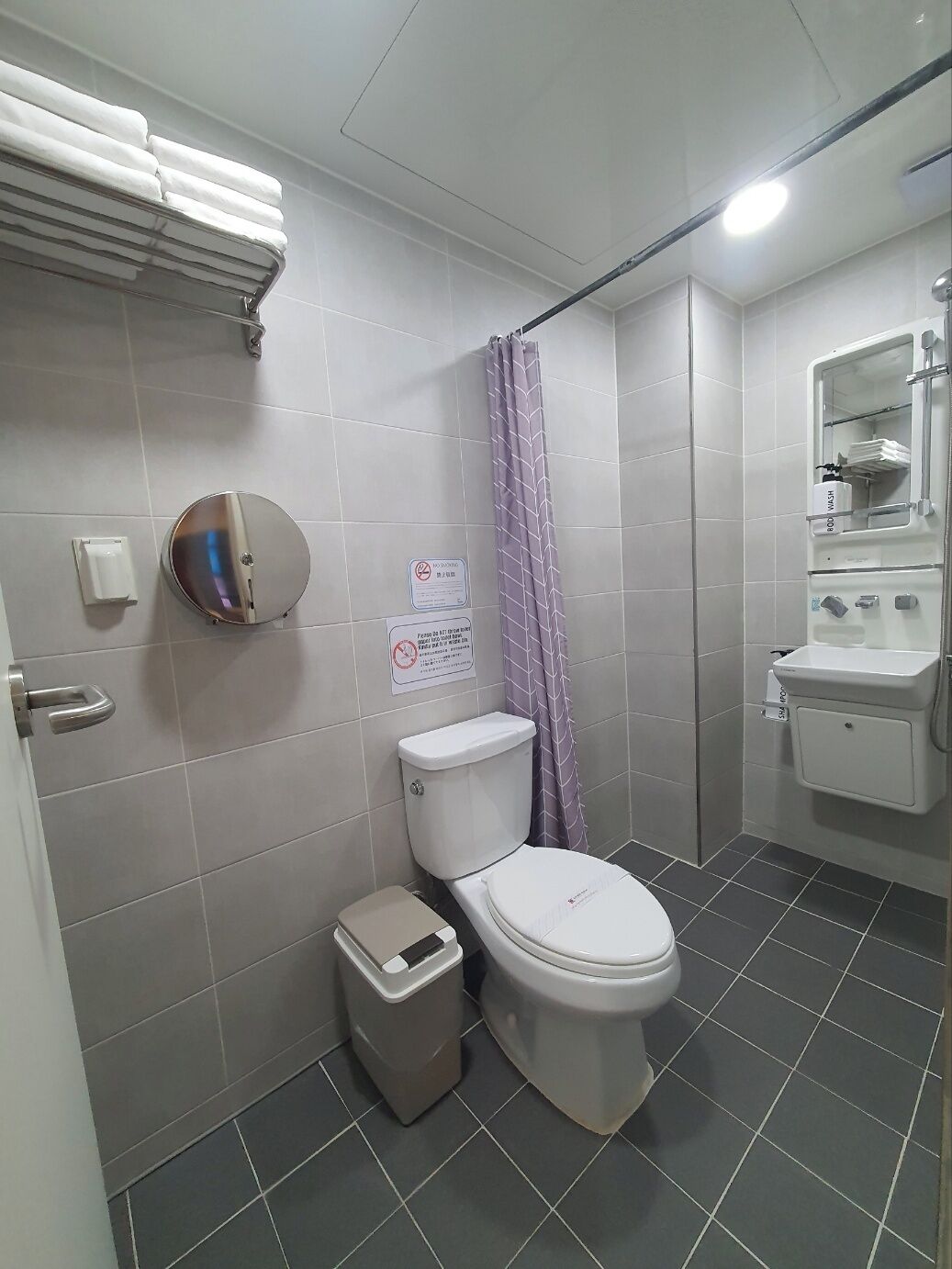 A bathroom with a toilet, a trash bin, a towel rack, a shower curtain, and a sink with a mirror.