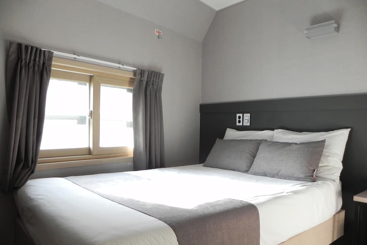 A modern hotel room with a large bed, grey curtains, a window with blinds, and a simple headboard.