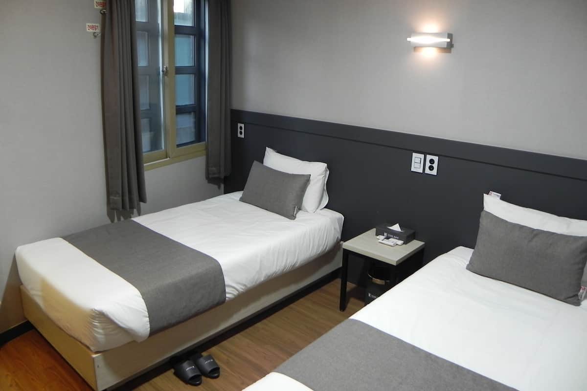 A hotel room with two beds, a window with curtains, a nightstand with a lamp, and a trash bin.