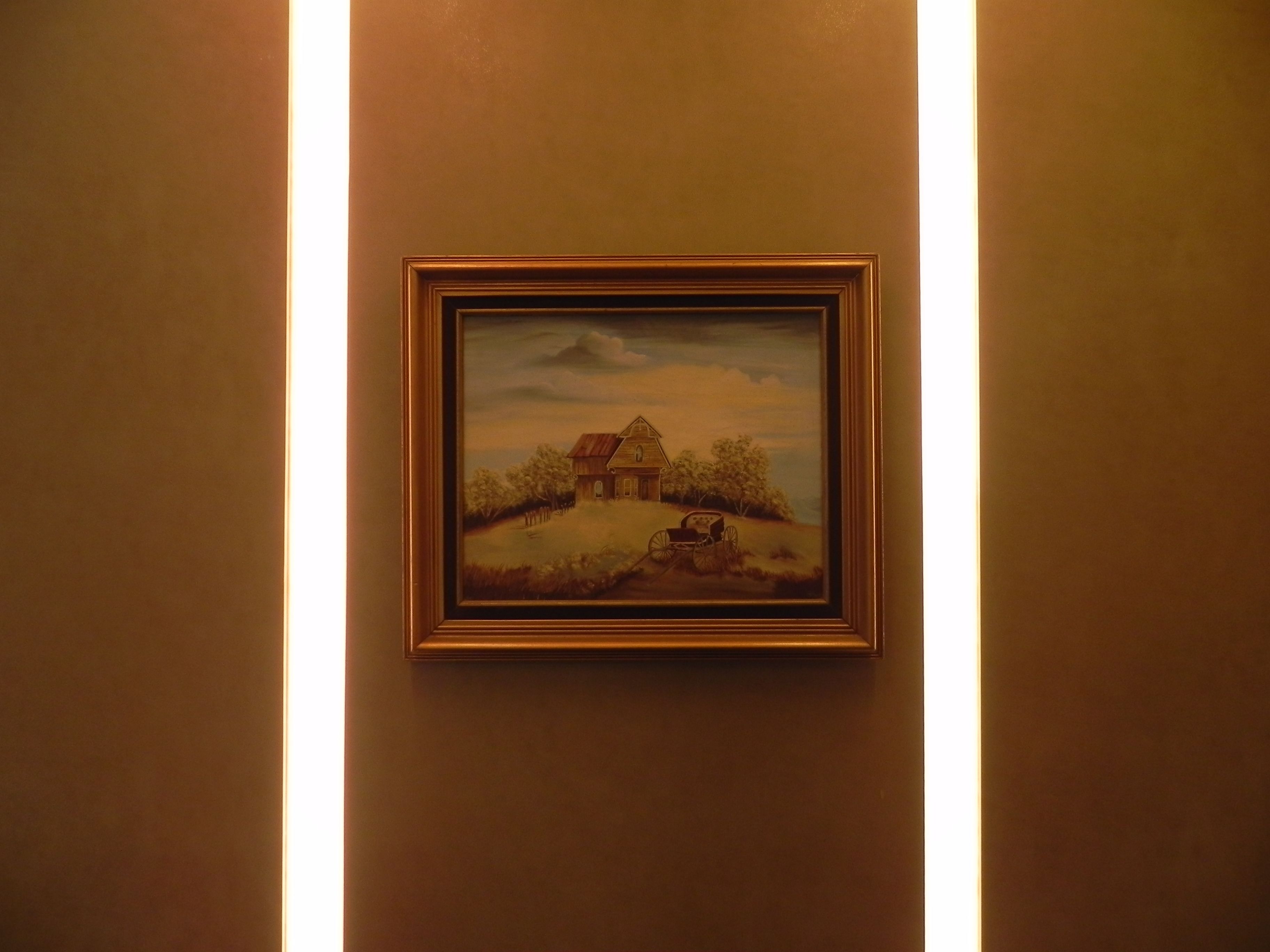 A framed painting of a rural scene with a house and a cart on a hill, mounted on a wall.