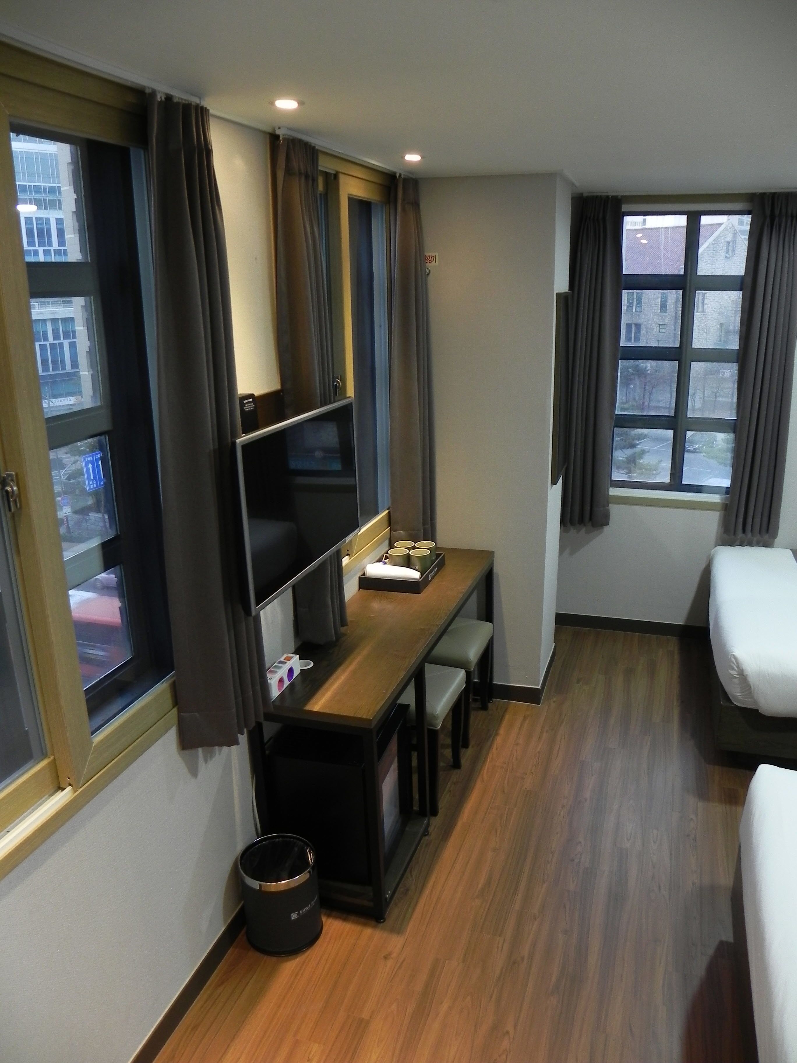 A compact hotel room with a wooden desk, a small seating area, a single bed, and a window with curtains.