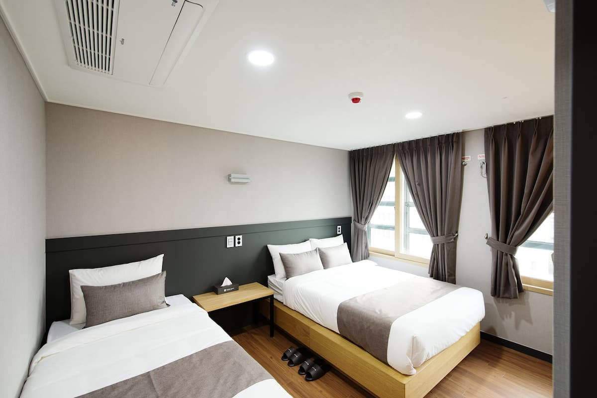 A hotel room with two beds, a window with curtains, a nightstand, and a wall-mounted air conditioning unit.