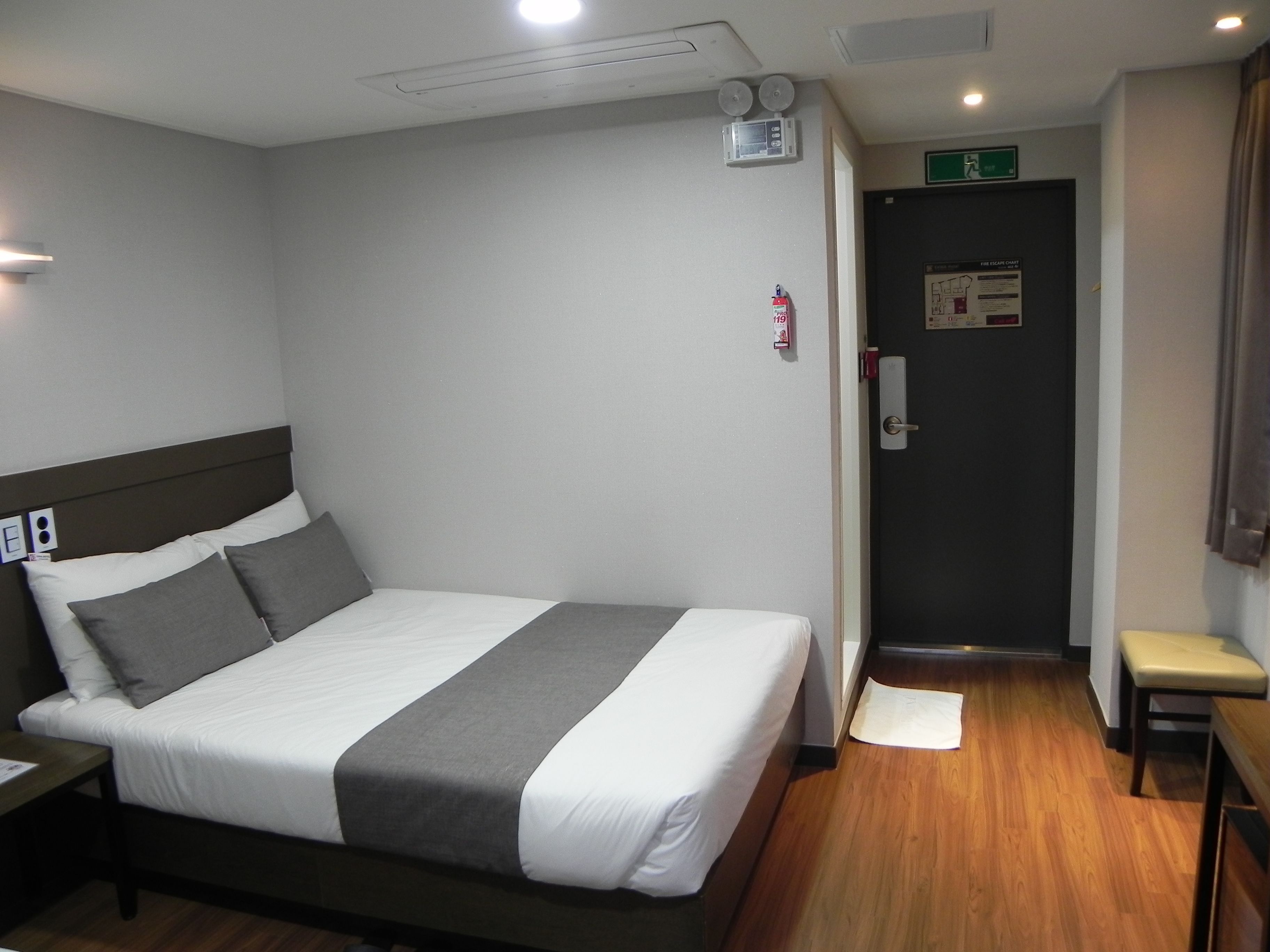 A hotel room with a bed, bedside table, and a small bench. There is a door with an emergency exit sign and a fire extinguisher.