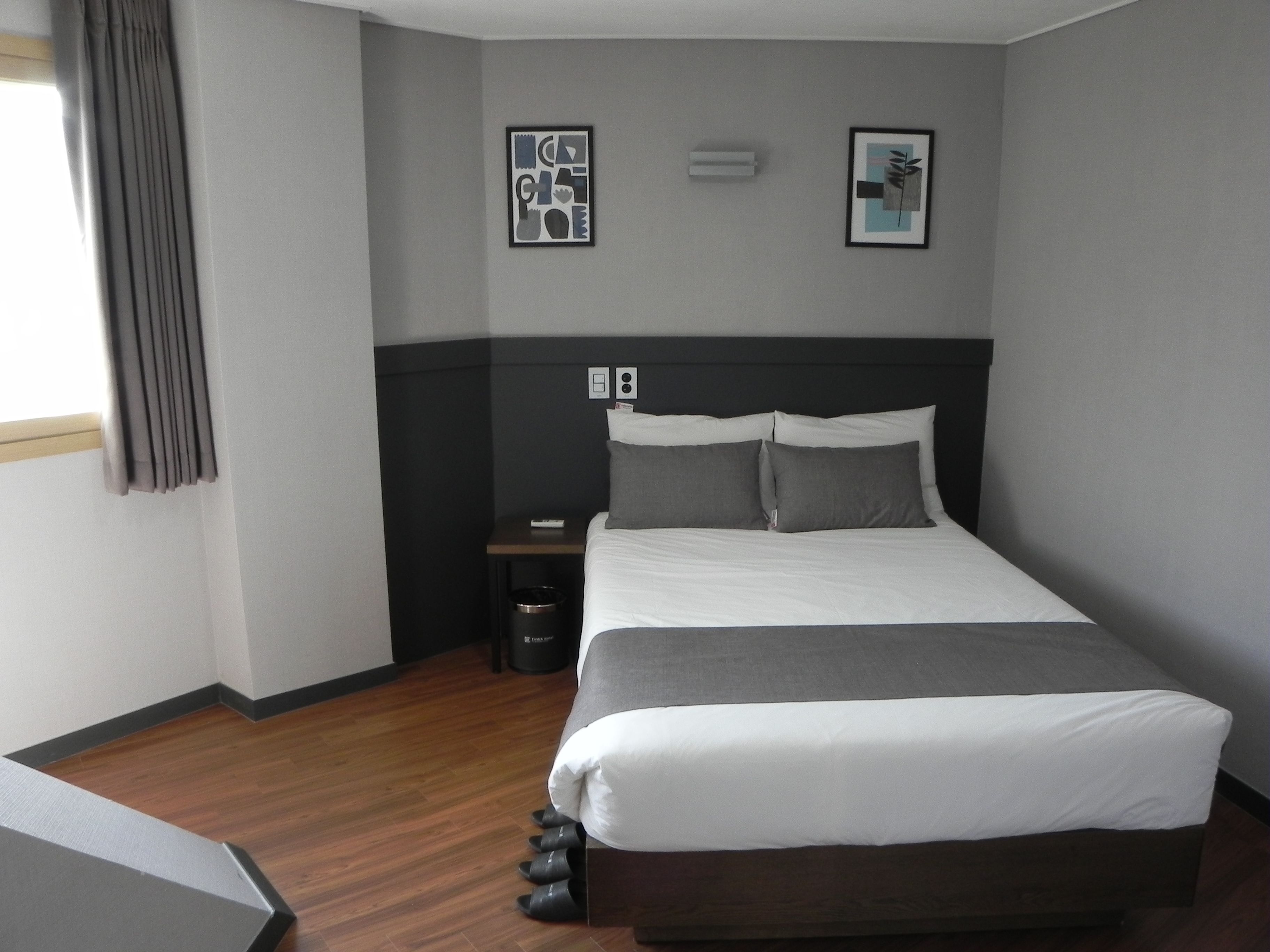 A modern hotel room with a large bed, a bedside table, a trash bin, and two framed pictures on the wall.