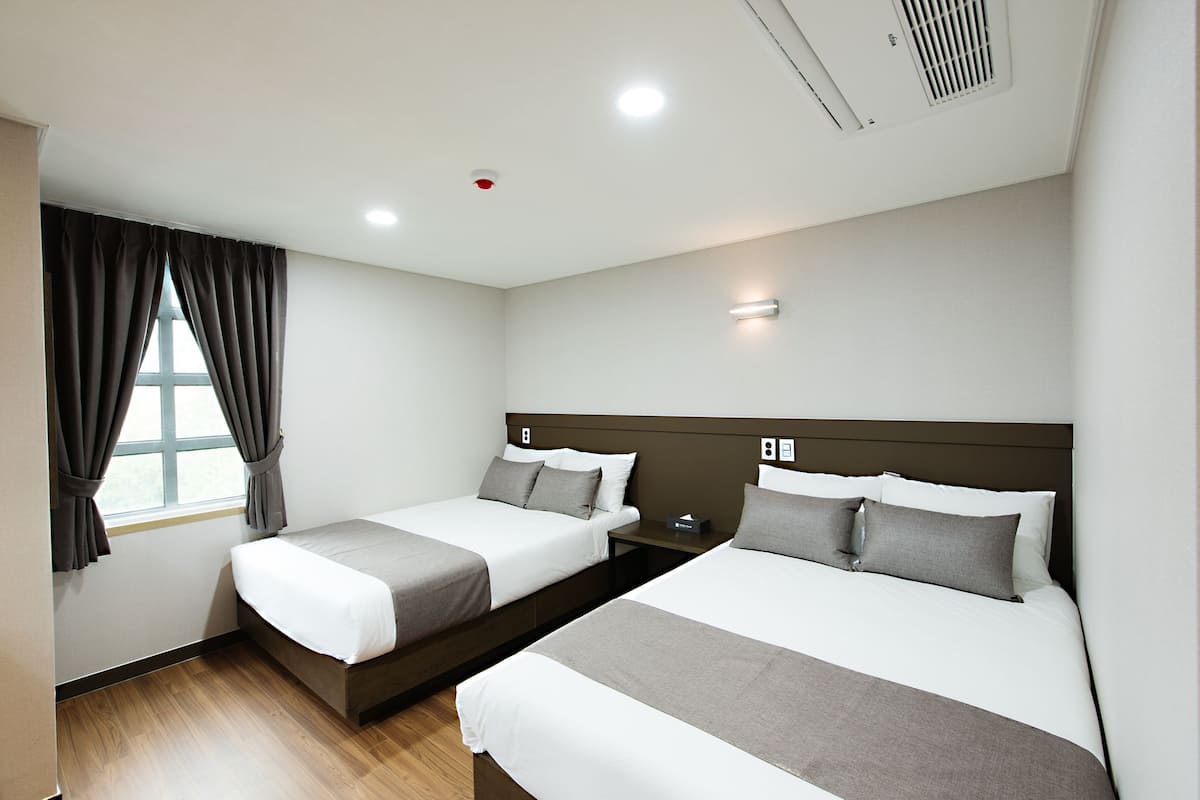 A hotel room with two beds, a window with curtains, a nightstand, and a wall-mounted light.
